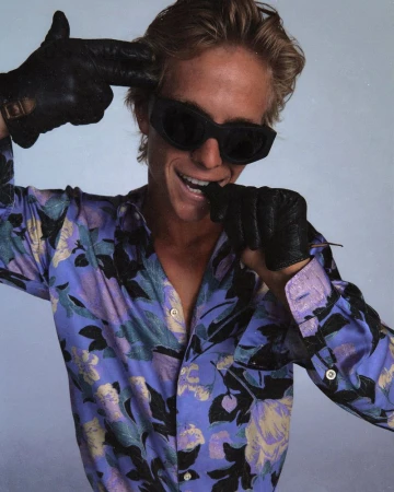A blonde adult man with dark sunglasses wears a patterned shirt and black gloves, with his fingers pointed at his head as if a gun.