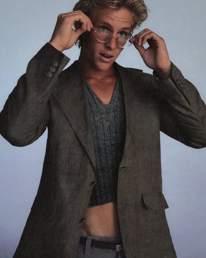 Blonde male model wearing round wire-rimmed glasses, a brown blazer, and grey v-neck top against a grey studio backdrop.