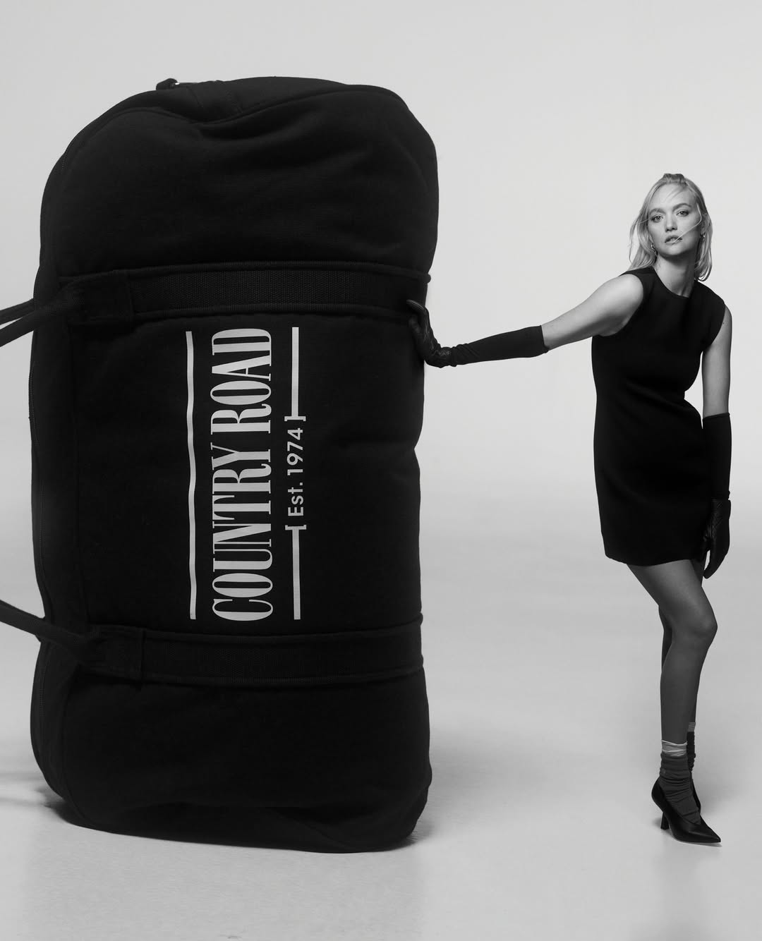 A woman in a black dress and gloves stands next to a large black Country Road duffel bag against a gradient white background in studio.