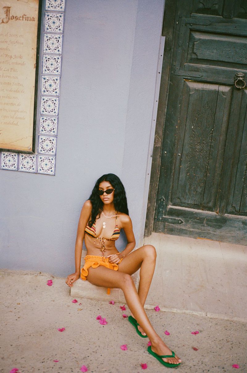 Woman wearing a bikini and skirt sitting on steps, accessorized with dark glasses and flip-flops, set against a wall with pink petals.