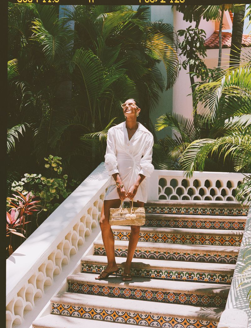 Woman in white dress and sunglasses poses on patterned steps, holding a handbag amid lush greenery, capturing a tropical, sunlit fashion moment.