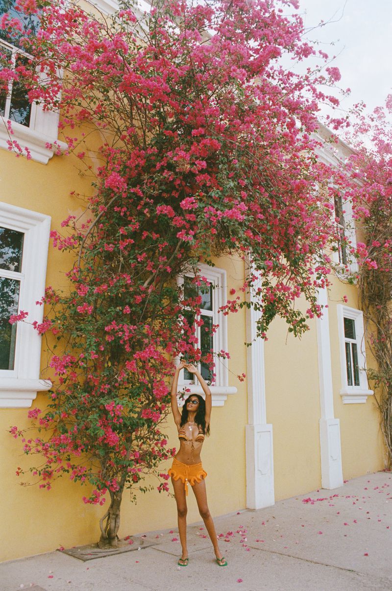 Adult woman poses in front of a yellow building, adorned with bright pink flowers, wearing an orange two-piece swimsuit on a sunny day.
