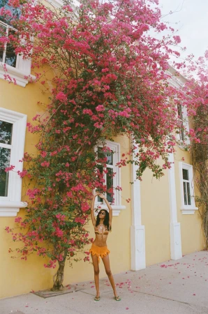 Adult woman poses in front of a yellow building, adorned with bright pink flowers, wearing an orange two-piece swimsuit on a sunny day.
