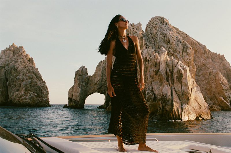 Woman in crochet halter dress standing on yacht against rock formation and ocean backdrop, wearing necklace and sunglasses.