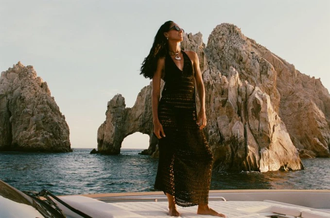 Woman in crochet halter dress standing on yacht against rock formation and ocean backdrop, wearing necklace and sunglasses.