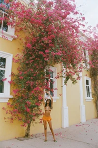 Model posing in an orange ruffled mini skirt and bikini top beneath a blooming pink bougainvillea tree against a yellow building.