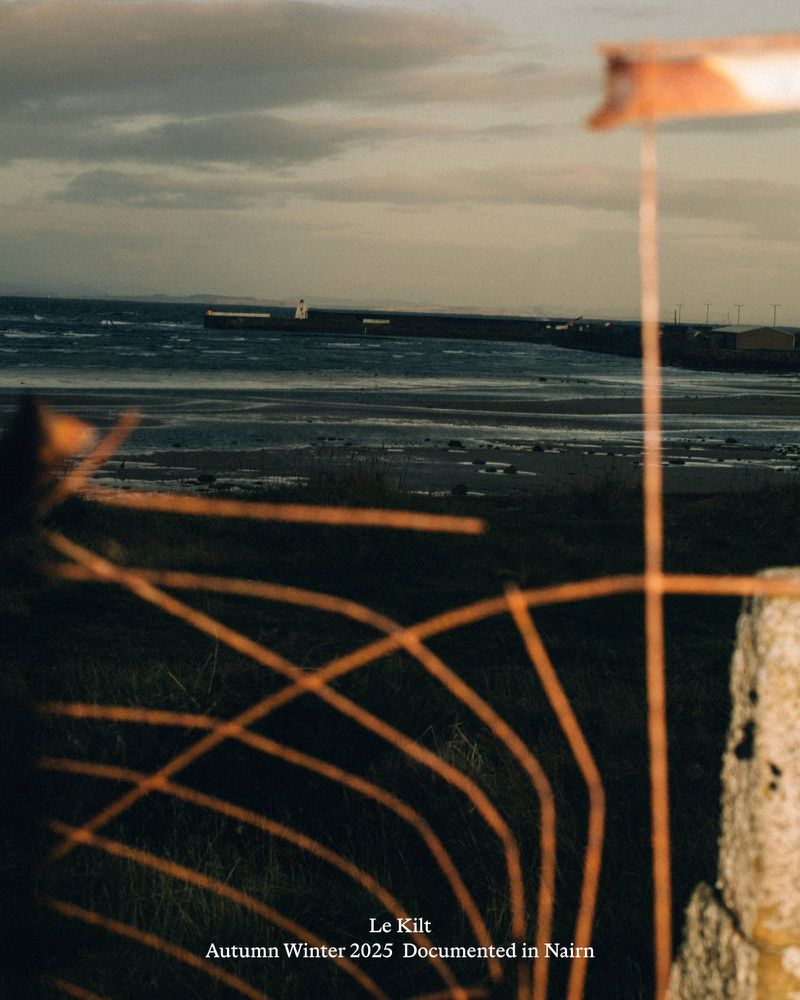 A landscape image of a body of water with a breakwater, a grassy field, and a fence in the foreground under a cloudy sky.
