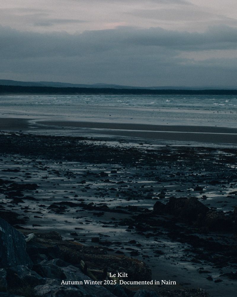 Moody coastal view with a muted color palette, featuring a beach at low tide, distant land, and cloudy sky. Rocks in the foreground.