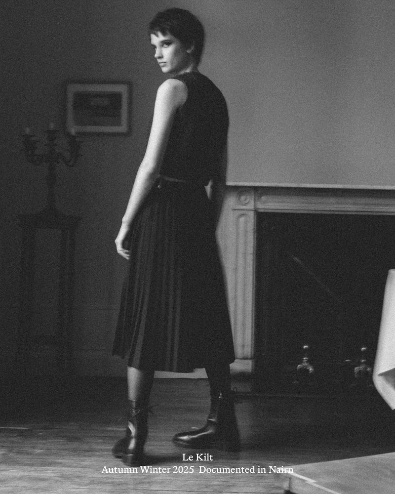 Monochrome fashion portrait of a woman in a pleated skirt and boots, gazing over her shoulder in an elegant indoor setting.