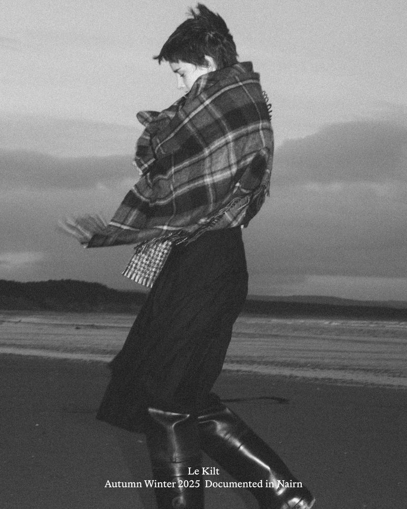 A person in a kilt and plaid shawl is walking on a beach with hills in the background, capturing a fashion moment in a grayscale photograph.