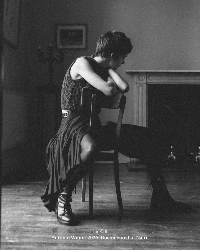 A person with short hair sits on a chair in a monochromatic indoor shot, wearing a knitted top, dark skirt, and boots, leaning forward.