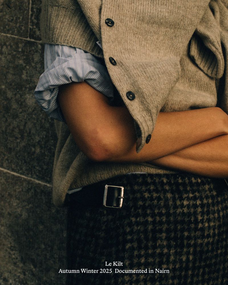 Close-up of a person wearing a beige cardigan, striped shirt, and a grey, patterned kilt, with arms crossed and a stone wall background.