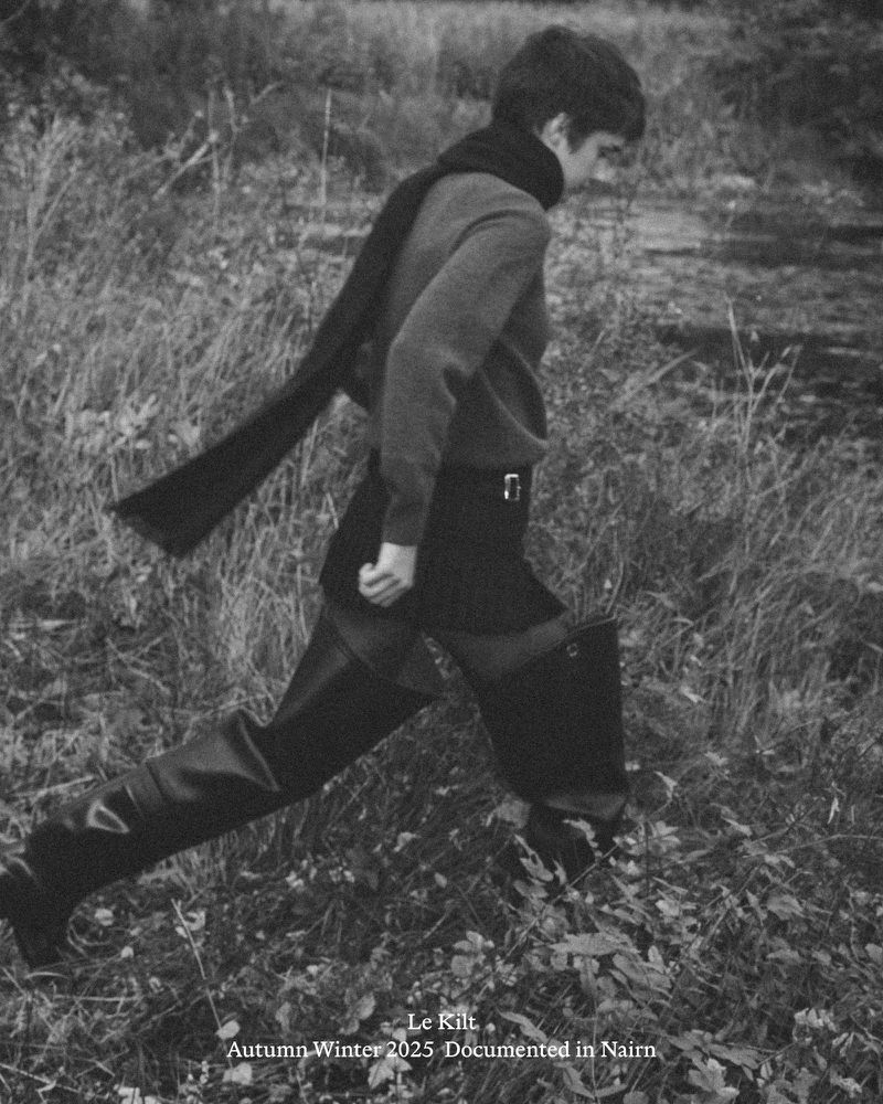 Monochrome outdoor shot of a person wearing a kilt, scarf, sweater, and boots striding across a grassy field with trees in the background.