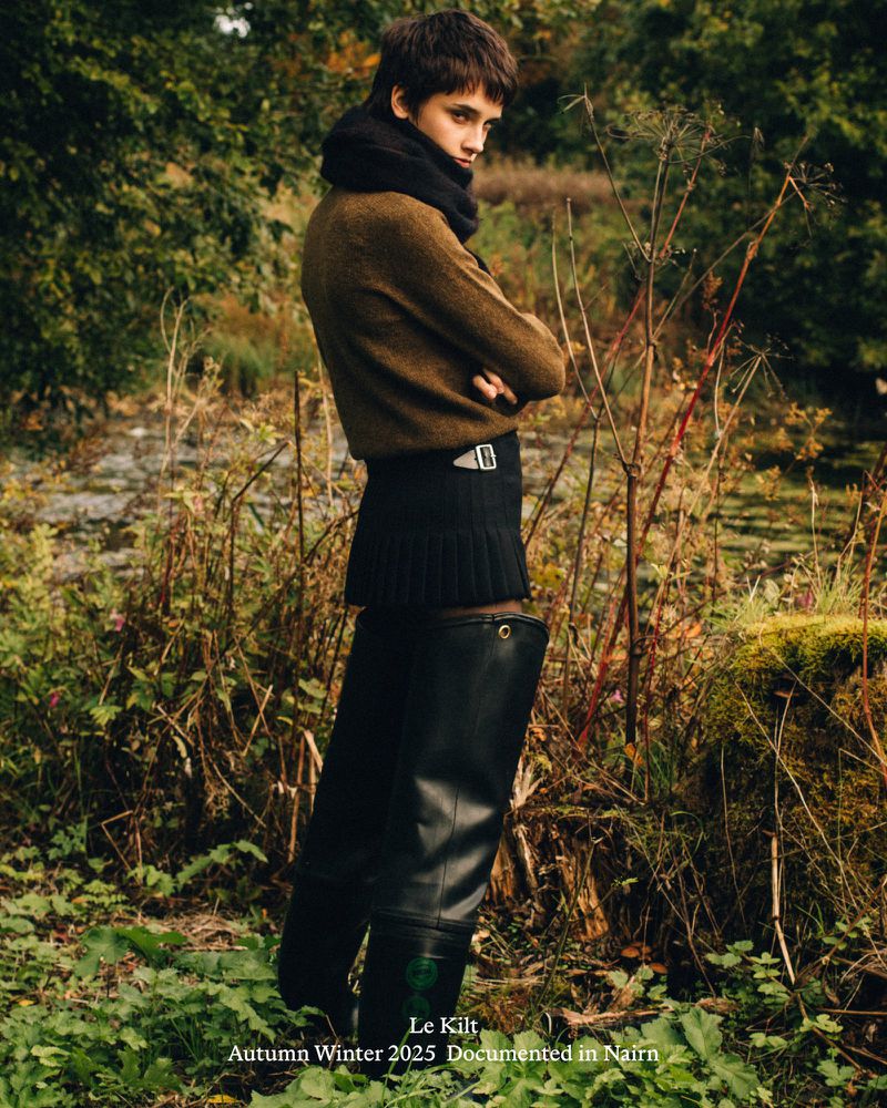 A person stands in a wooded landscape wearing a brown sweater, kilt, black scarf, and black rubber boots, looking off to the side.