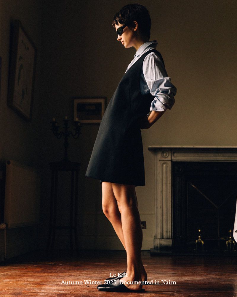 Fashion editorial shot of a woman in a blue dress shirt, black pinafore dress, and sunglasses in a room with dark wood floors.