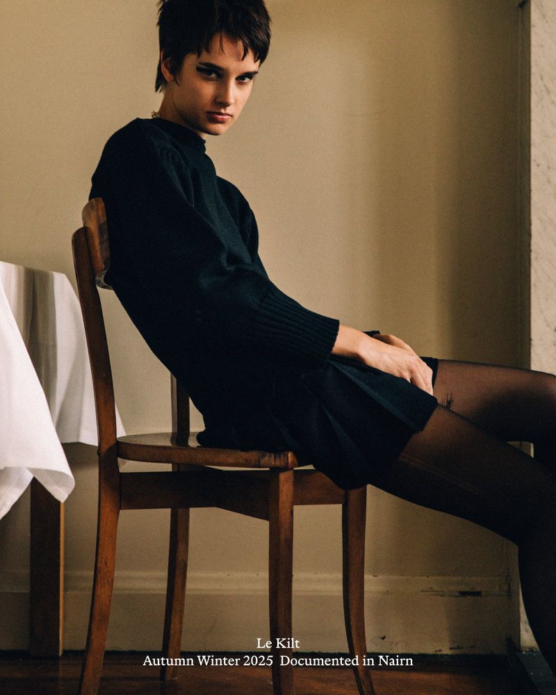 A woman with short dark hair, wearing a black dress and tights, sits sideways on a wooden chair in a softly lit indoor setting.