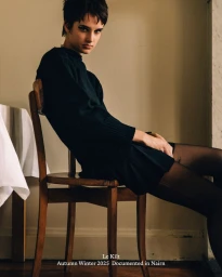 Model with short pixie hair wearing an all-black long-sleeved knit dress and tights seated on a wooden chair.