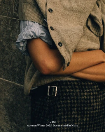Close-up of a person with crossed arms wearing a beige knit cardigan and a houndstooth kilt with a metal buckle.