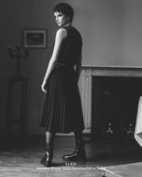 Model with short hair looking over her shoulder wearing a black sleeveless pleated dress and leather boots beside a fireplace.