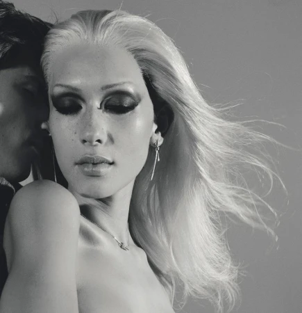 Close-up black and white portrait of a blonde model with dramatic dark eye makeup posing next to another person.