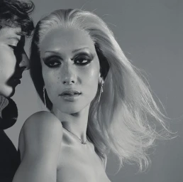 Portrait of a blonde woman with heavy eye makeup and a profile silhouette of a man against a plain grey background