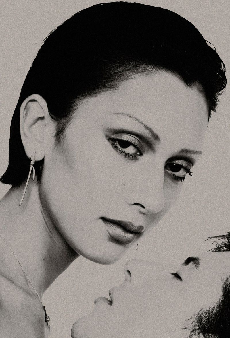 Close-up monochrome portrait of two people with slicked-back dark hair and dramatic makeup against a light background.