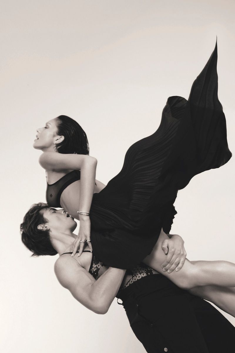 Two women in a dynamic pose: One woman holding the other, who is wearing a black dress and facing upwards against a bright background.