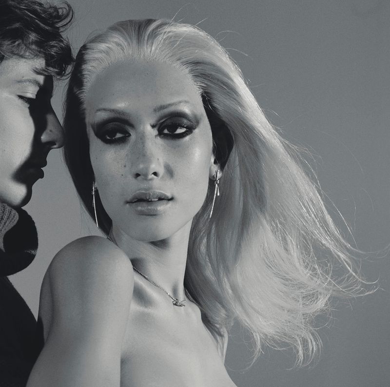 Striking monochrome portrait of a fair-haired woman with heavy makeup, complemented by a partial view of another person with short dark hair.