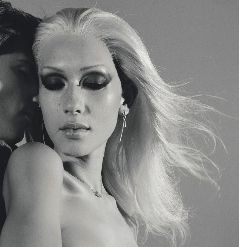 Monochrome portrait of a fair-skinned woman with a man, heavy eye makeup, and flowing hair, wearing jewelry, against a plain background.