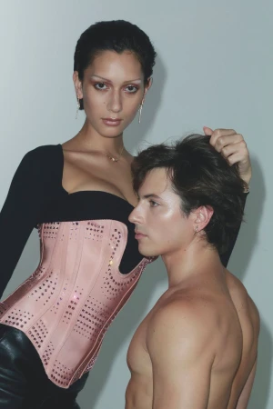 A model in a pink corset places a hand on another shirtless model's head against a white backdrop in a fashion forward studio shot.