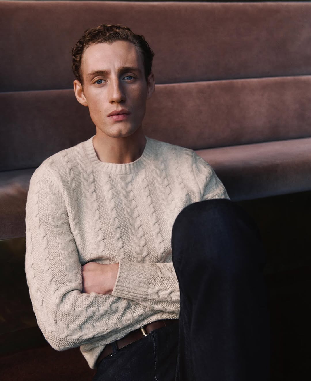Indoor portrait of a young adult male with light skin and curly brown hair wearing a cream-colored cable knit sweater and dark pants.