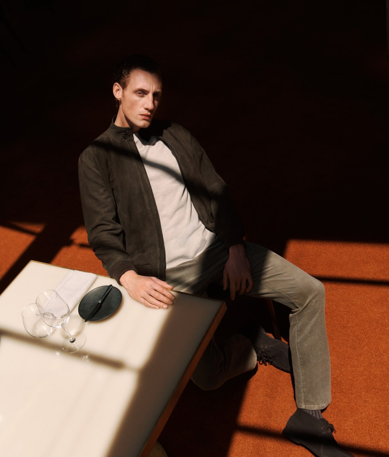 Male model poses indoors with natural light, wearing a jacket and trousers, sitting next to a table with tableware, editorial fashion photography.