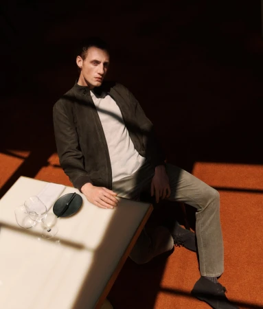 Man seated next to a table with wine glasses wearing a dark jacket and trousers in dramatic lighting for Trenery