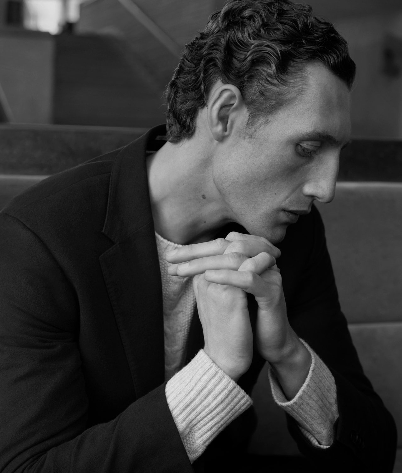 Portrait of a man sitting on stairs in a blazer and sweater with hands clasped, black and white photography.