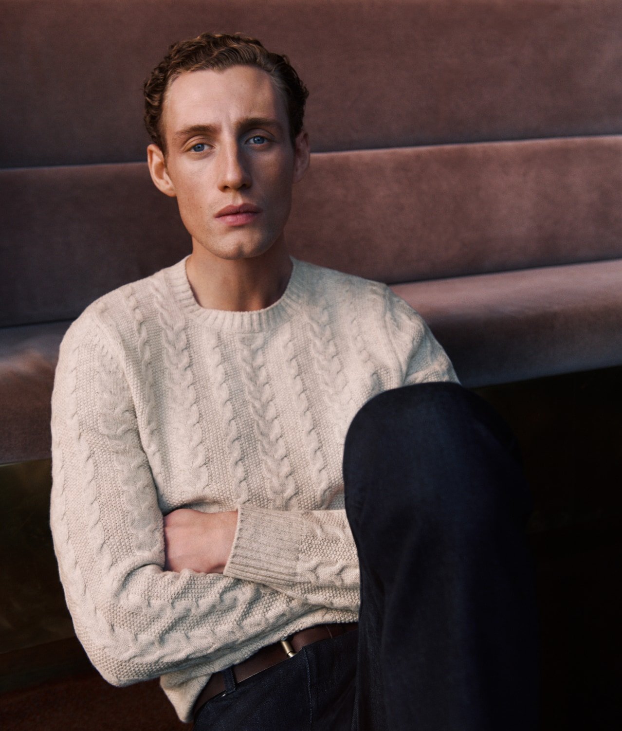 Portrait of a young adult male sitting with his arms crossed while wearing a sweater and dark pants in a casual pose indoors.