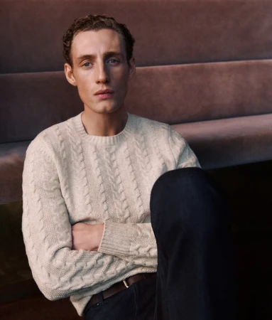 Portrait of a young adult male sitting with his arms crossed while wearing a sweater and dark pants in a casual pose indoors.