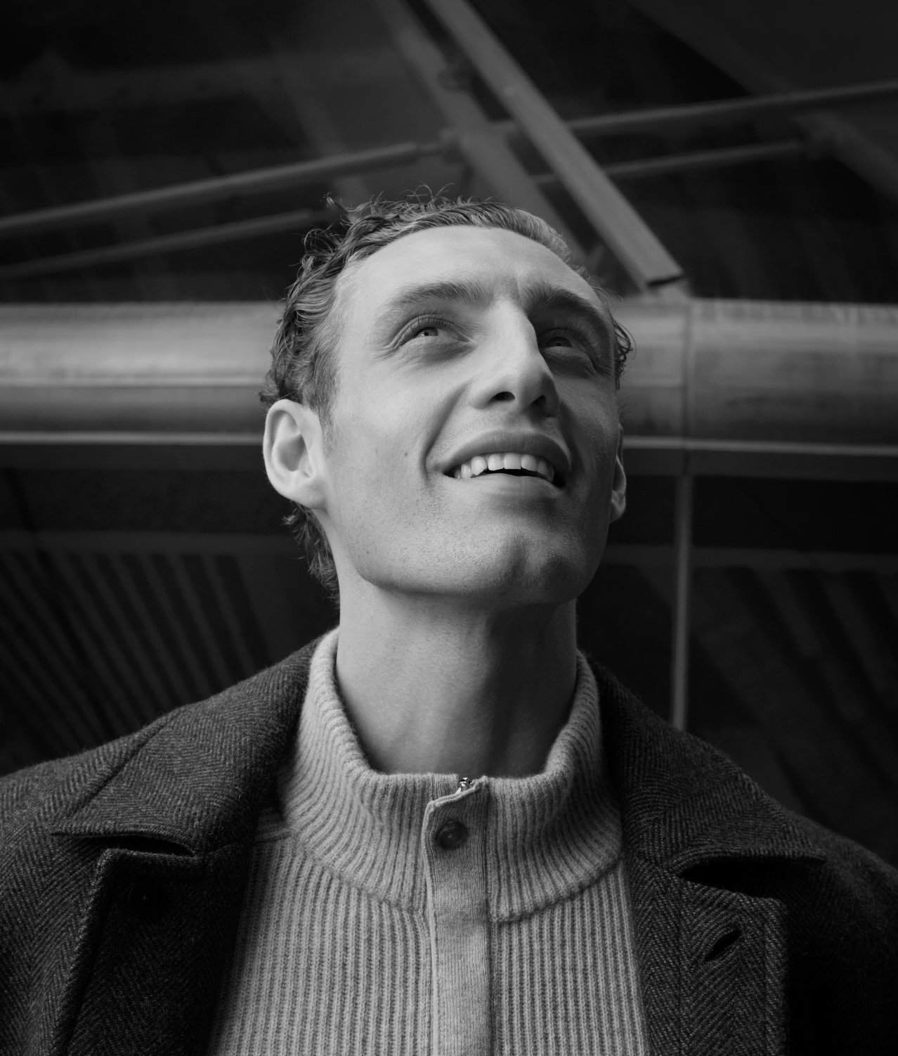 Black and white portrait of smiling adult male in jacket looking up against structural background.