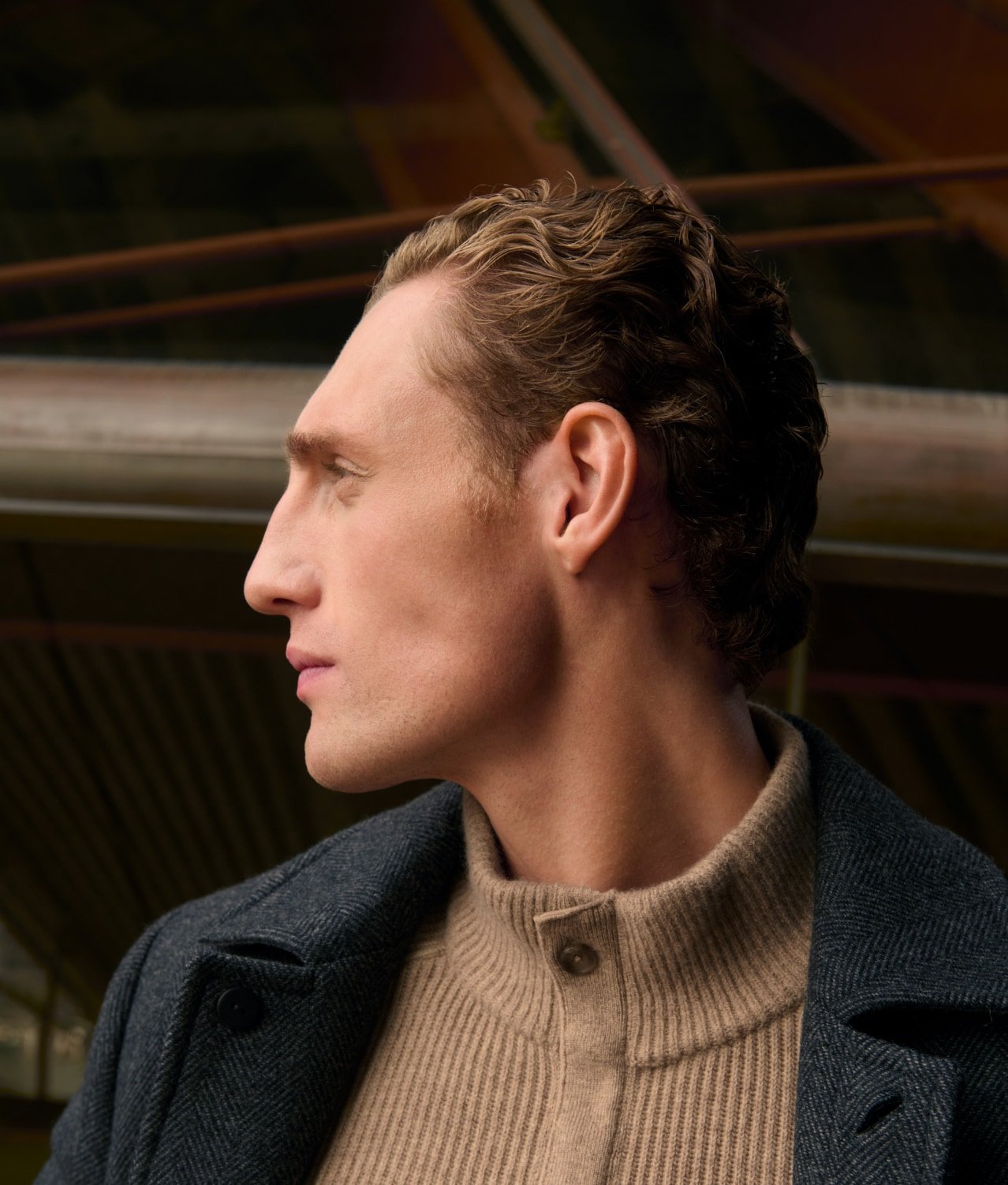 Side profile portrait of an adult male with blonde hair, wearing a beige sweater and a grey coat against a blurry background.