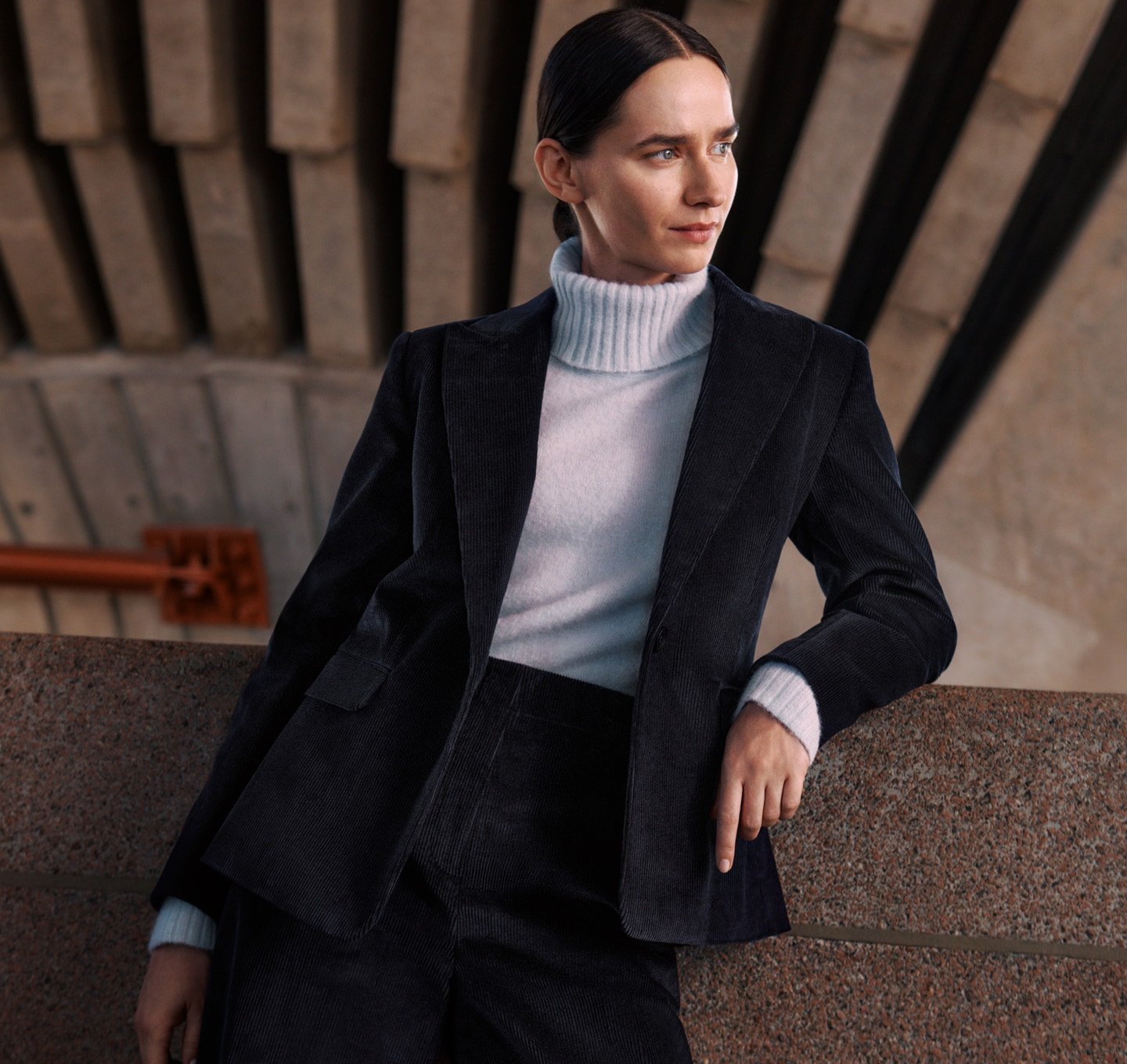 A woman in a stylish corduroy suit and a light blue turtleneck sweater poses against an architectural wall, looking off to the side.