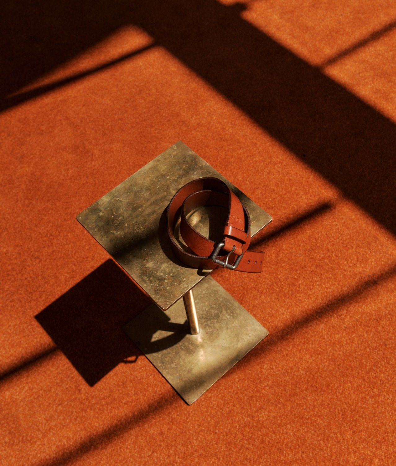 Tan leather belt with a metal buckle resting on a gold metal accent table, placed on an orange textured carpet, with light and shadows.