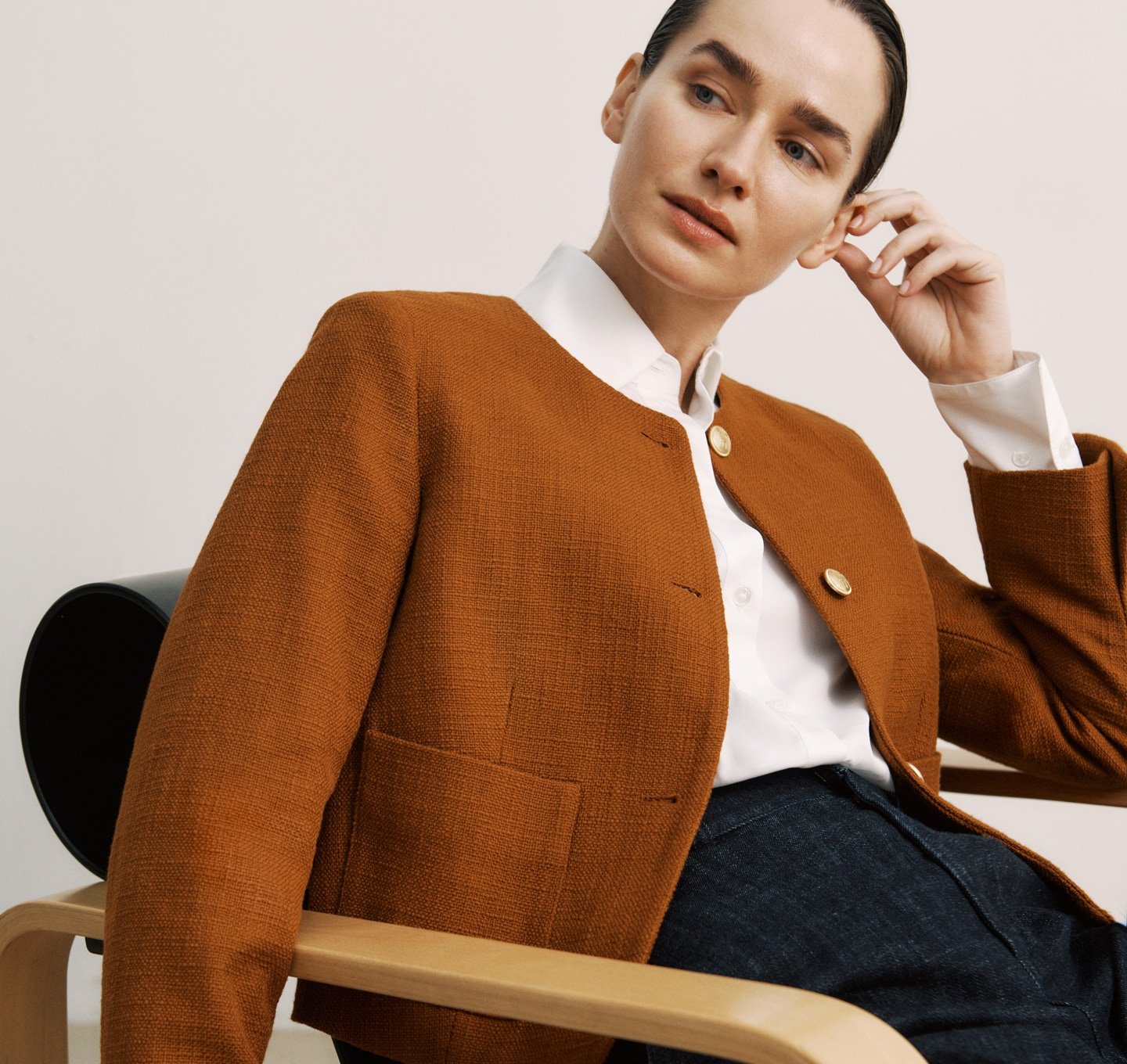 A woman in a brown jacket and white shirt sits in a wooden chair against a white background in a fashion portrait.
