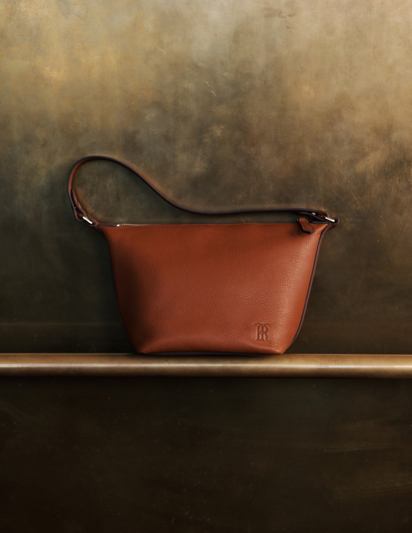 Brown leather handbag with a short handle, displayed on a gold bar against a textured brown and gold backdrop, emphasizing its design.