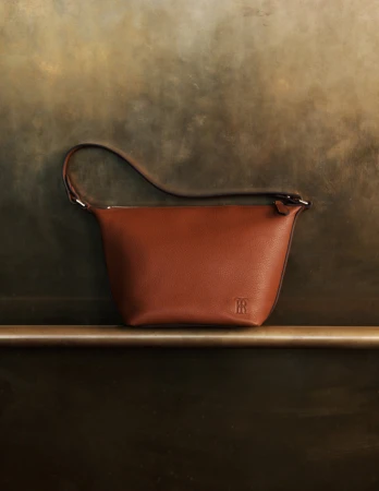 Brown leather handbag with a short handle, displayed on a gold bar against a textured brown and gold backdrop, emphasizing its design.