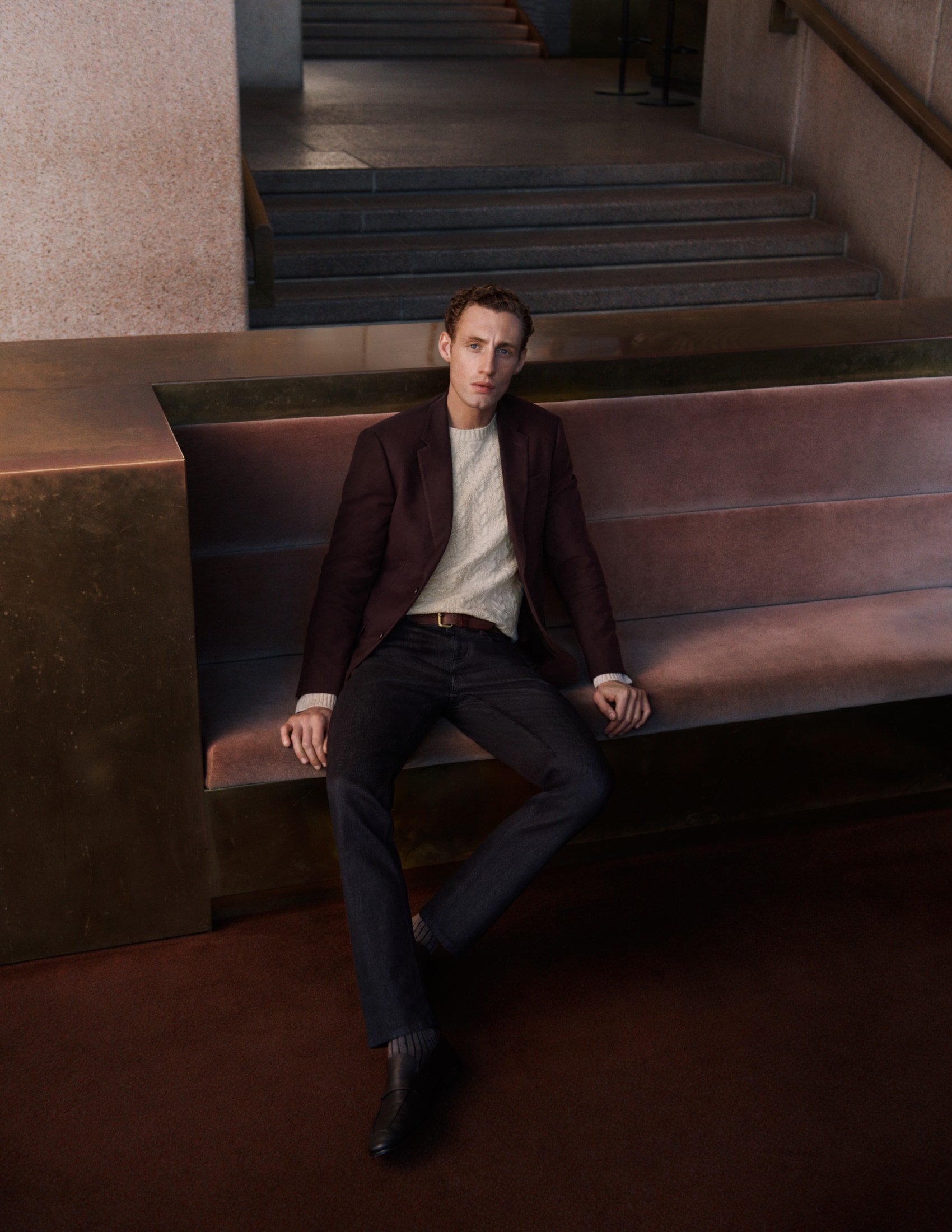 An adult male sits on a bench wearing a maroon blazer, light-colored sweater, dark pants and shoes, in a modern indoor setting.
