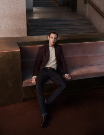 An adult male sits on a bench wearing a maroon blazer, light-colored sweater, dark pants and shoes, in a modern indoor setting.