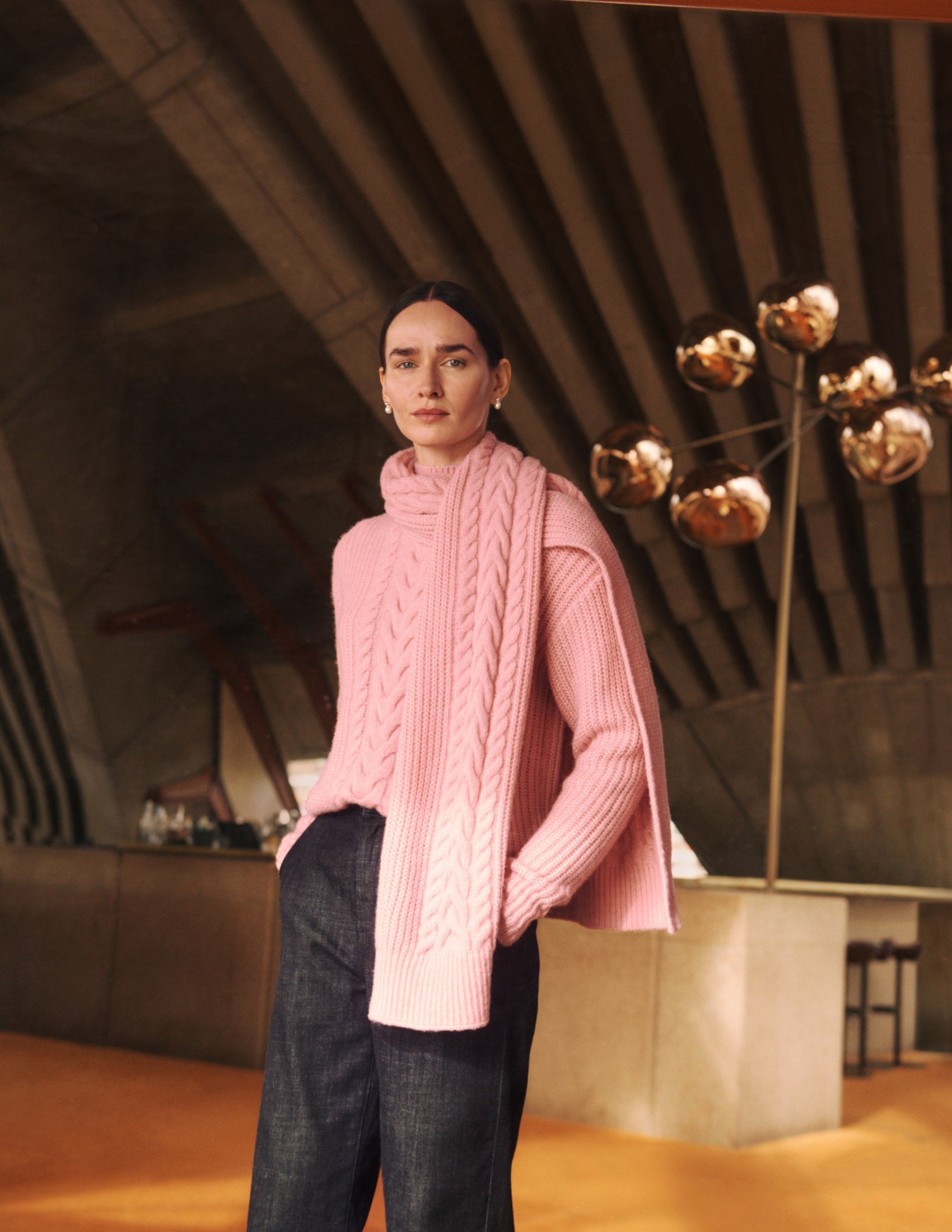 Adult woman poses in a pink cable knit sweater and dark pants against an architectural backdrop with globe lighting.