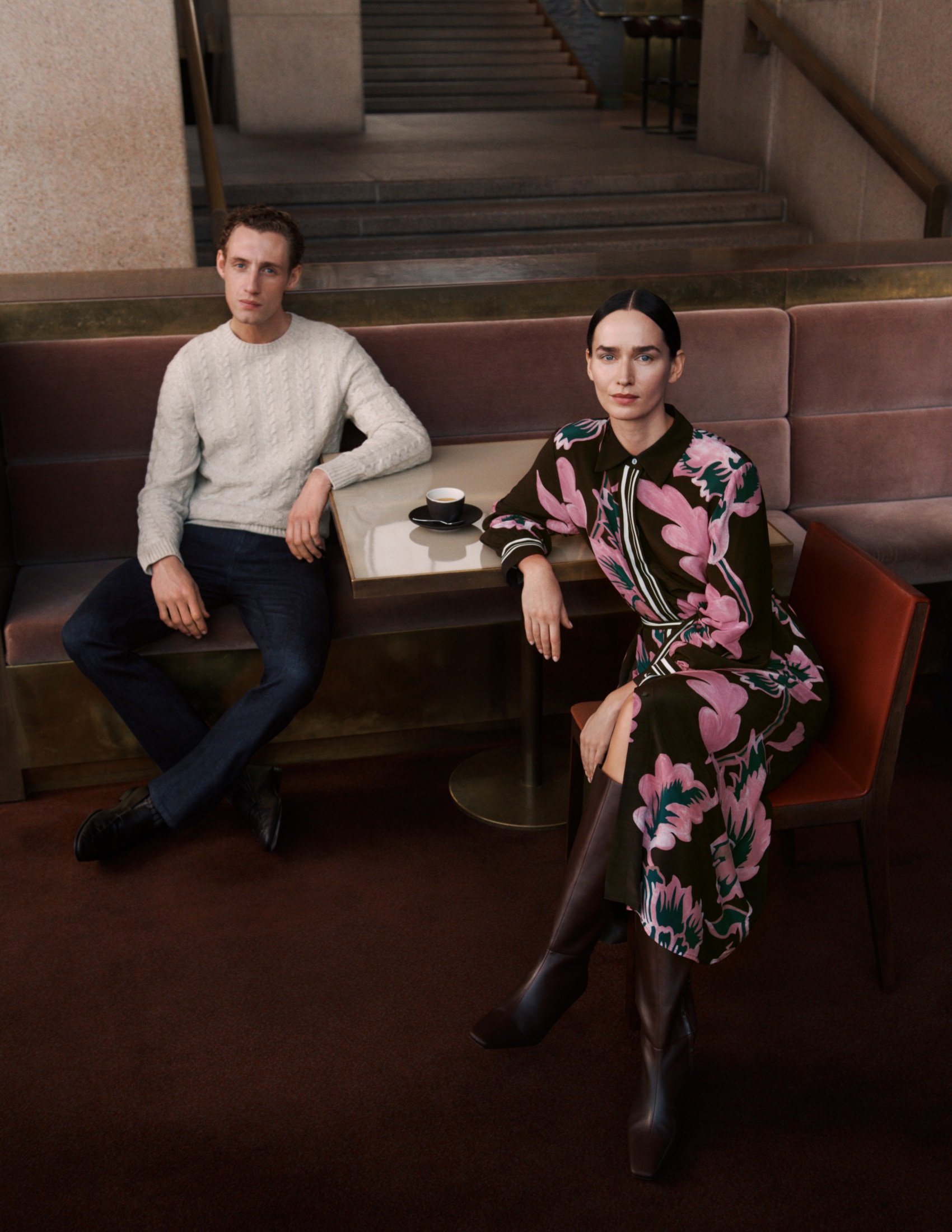 Elegant couple seated in a cafe: woman in floral dress and boots, man in sweater and jeans, creating a stylish indoor scene.