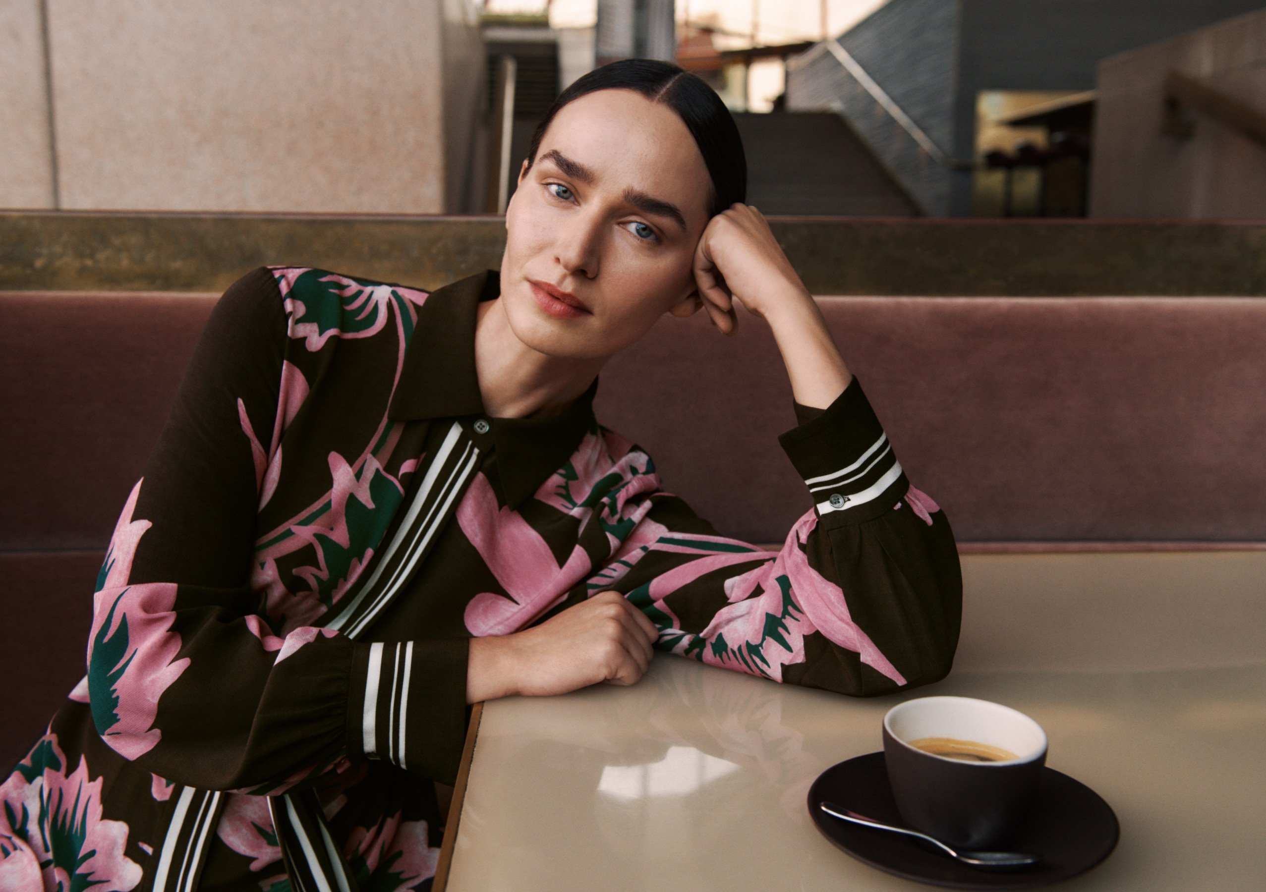 Adult woman in a floral print blouse sitting at a table next to a cup of coffee in a cafe setting, looking at the camera.