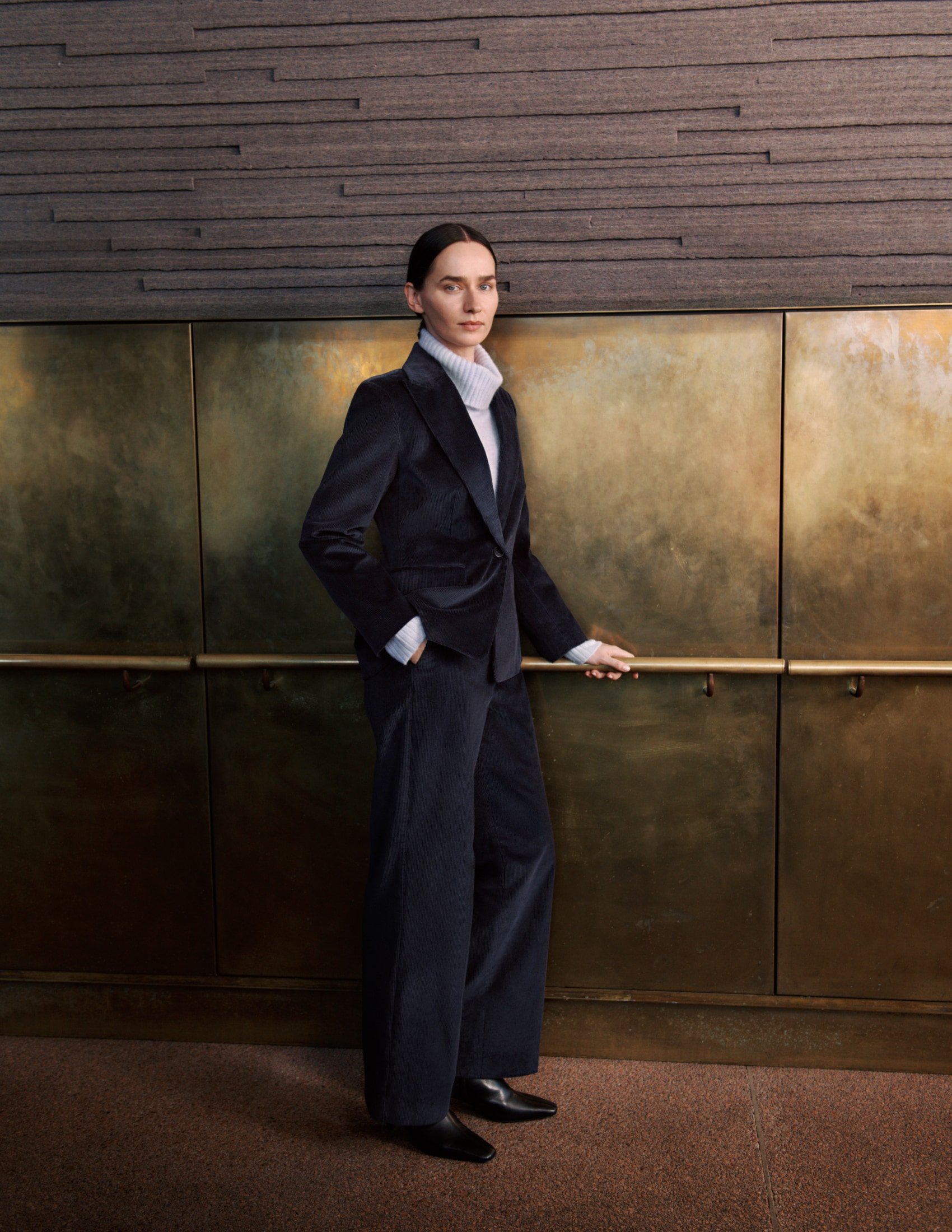 Adult female in a dark blue velvet suit with a white turtleneck poses against a textured wall with a vintage brass elevator.