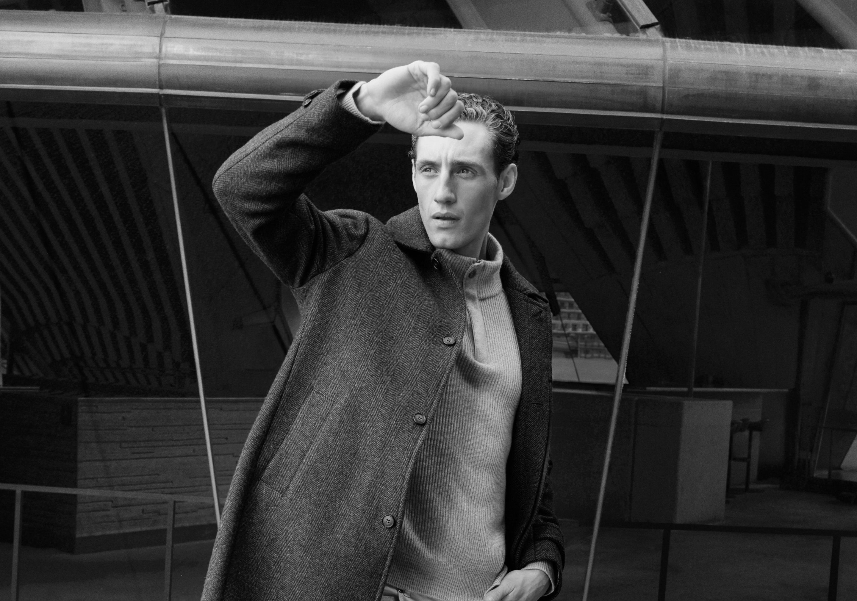 Man in a wool coat shielding his eyes, standing outside with neutral expression in black and white, looking into the distance.